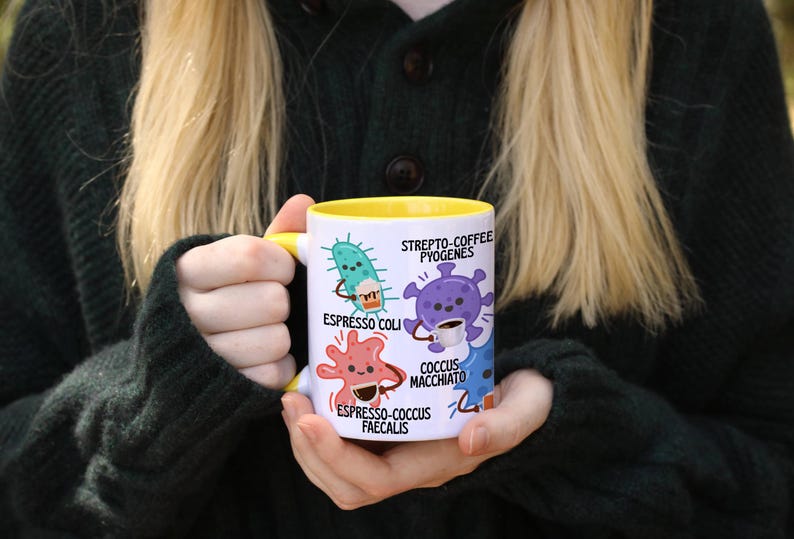 May include: A white ceramic mug with a yellow interior and handle, featuring cartoon illustrations of bacteria. The mug has the text "Espresso Coli", "Strepto-Coffee Pyogenes", "Coccus Macchiato", and "Espresso-Coccus Faecalis".