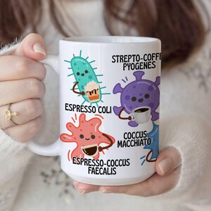 May include: White ceramic coffee mug featuring cartoon microbes with coffee-themed names like "Espresso Coli" and "Coccus Macchiato." The mug has a large handle and is held by a person wearing a white sweater. The mug is a humorous gift idea.