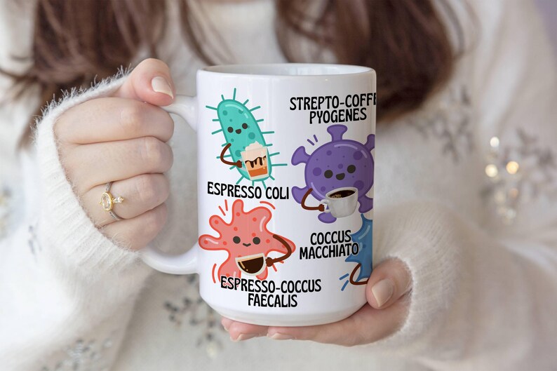 May include: White ceramic coffee mug featuring cartoon microbes with coffee-themed names like "Espresso Coli" and "Coccus Macchiato." The mug has a large handle and is held by a person wearing a white sweater. The mug is a humorous gift idea.