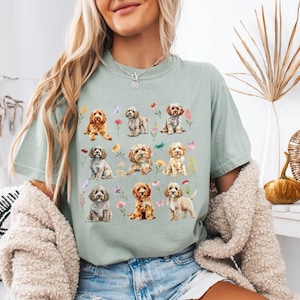 May include: A light green t-shirt featuring a watercolor design of various puppies with flowers and butterflies. The puppies are in different poses and colors, creating a charming and whimsical aesthetic. The shirt is a relaxed fit.
