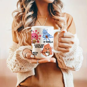 May include: White ceramic mug featuring cartoon illustrations of microbes with coffee-related puns. The mug has a handle and is held by a person. The text on the mug includes "SIP-MONELLA" and "CLOSTRI-DRIP BOTULINUM".
