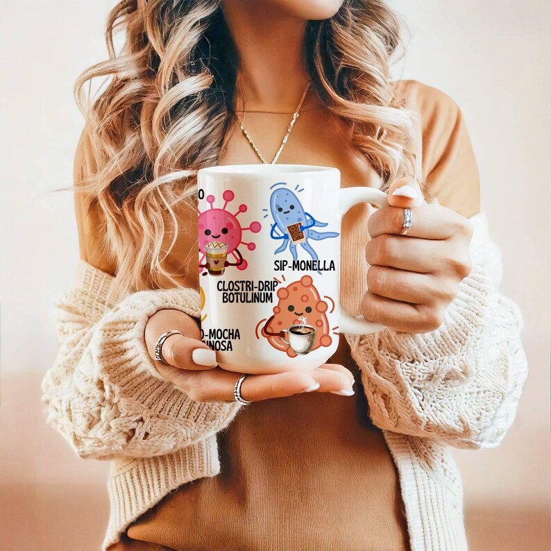 May include: White ceramic mug featuring cartoon illustrations of microbes with coffee-related puns. The mug has a handle and is held by a person. The text on the mug includes "SIP-MONELLA" and "CLOSTRI-DRIP BOTULINUM".