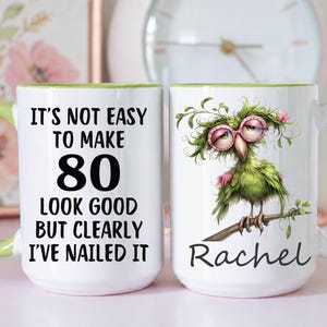 May include: A white ceramic mug with a light green handle and interior. The front features black text reading IT'S NOT EASY TO MAKE 80 LOOK GOOD BUT CLEARLY I'VE NAILED IT. The back shows a green bird wearing pink glasses and the name Rachel.