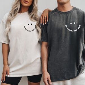 Könnte beinhalten: Zwei T-Shirts, eines in Creme und eines in Anthrazitgrau, mit einem Smiley-Gesicht-Design. Das cremefarbene Shirt hat den Text "Rachel Smith, DSW. 2025" und das graue Shirt hat "DR. SMITH. DSW. 2025" auf der Vorderseite aufgedruckt.