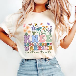 Könnte beinhalten: Cremefarbenes T-Shirt mit floralem Design und den Worten "KNEE REPLACEMENT WARRIOR" in bunten Buchstaben im Retro-Stil. Das Shirt enthält auch die Worte "custom text" darunter.