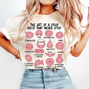 Könnte beinhalten: Cremefarbenes T-Shirt mit einer rosa Illustration verschiedener roter Blutkörperchenformen mit Gesichtern. Der Text "THE ART OF A DROP RBCS THAT NEVER STOP" ist über den Illustrationen aufgedruckt. Das T-Shirt wird von einer Person getragen.