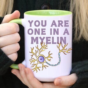 May include: White ceramic mug with a green handle and interior. The mug features the text "YOU ARE ONE IN A MYELIN" in purple, along with a yellow and blue neuron illustration. The mug is held by a person.