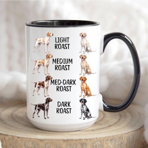 Puede incluir: Una taza de cerámica blanca con asa y borde interior negros. La taza presenta ilustraciones de perros con diversos colores de pelaje junto a las etiquetas: Light Roast, Medium Roast, Med-Dark Roast y Dark Roast, dispuestas en cuatro filas.