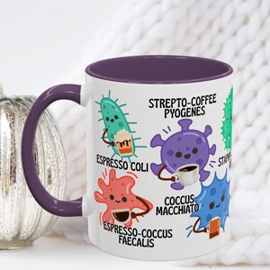 May include: White ceramic coffee mug with a purple interior and handle. The mug features cartoon illustrations of bacteria holding coffee cups, with text including "Espresso Coli" and "Coccus Macchiato".