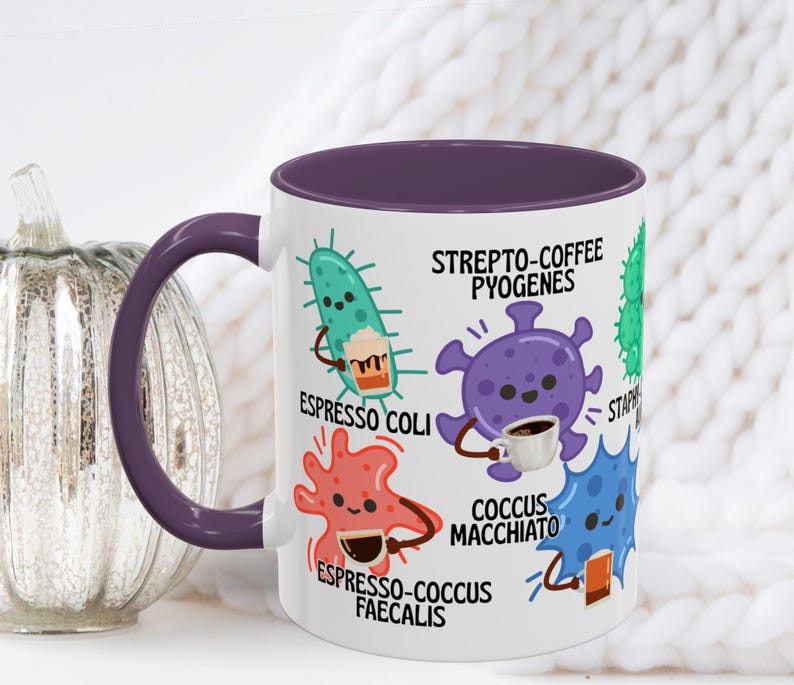 May include: White ceramic coffee mug with a purple interior and handle. The mug features cartoon illustrations of bacteria holding coffee cups, with text including "Espresso Coli" and "Coccus Macchiato".