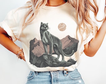 Könnte beinhalten: Cremefarbenes T-Shirt mit einer detaillierten Illustration eines Pumas, der auf einem Felsvorsprung mit Bergen und einer Sonne steht. Der Puma ist in Grau- und Schwarztönen gehalten, die Berge in Brauntönen.