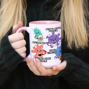 May include: White ceramic mug with a pink handle and interior, featuring cartoon illustrations of bacteria with coffee-themed names like "Espresso Coli" and "Coccus Macchiato." The mug is held by a person wearing a dark green sweater.