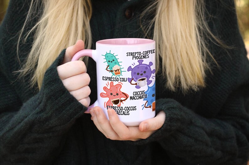 May include: White ceramic mug with a pink handle and interior, featuring cartoon illustrations of bacteria with coffee-themed names like "Espresso Coli" and "Coccus Macchiato." The mug is held by a person wearing a dark green sweater.