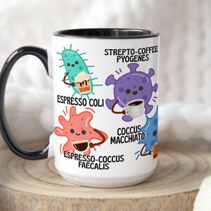 May include: White ceramic coffee mug with a black handle and rim. The mug features cartoon illustrations of bacteria, each holding a coffee cup. Text includes "Espresso Coli," "Strepto-Coffee Pyogenes," "Espresso-Cocus Faecalis," and "Cocus Macchiato."