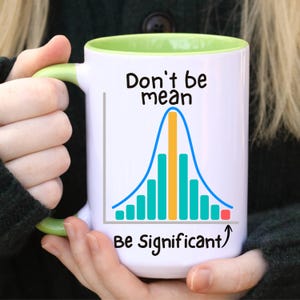 May include: White ceramic mug with a green handle and interior. The mug features the text "Don't be mean Be Significant" above a bell curve graph in blue, teal, and yellow.