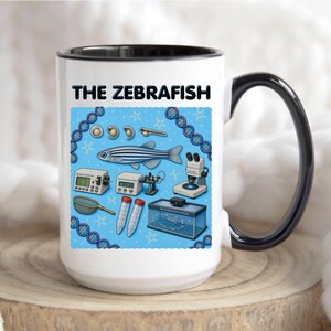 May include: A white ceramic mug with a black handle and interior rim, featuring a graphic print titled THE ZEBRAFISH. The design includes illustrations of zebrafish developmental stages, a microscope, lab equipment, and DNA strands on a blue background.