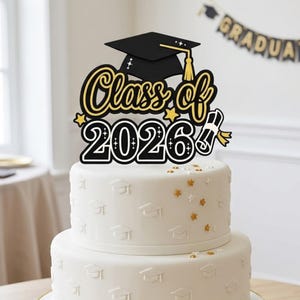 Op de afbeelding: Een witte taart met twee lagen en een zwarte en gouden taarttopper. De topper leest "Class of 2026" en heeft een afstudeerhoed. De taart is versierd met reliëf afstudeerhoeden en gouden sterrenstrooisel.