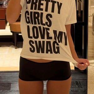 Puede incluir: Camiseta blanca con el texto "PRETTY GIRLS LOVE MY SWAG" en letras negras. La camiseta se combina con pantalones cortos negros. La imagen está tomada en interiores.