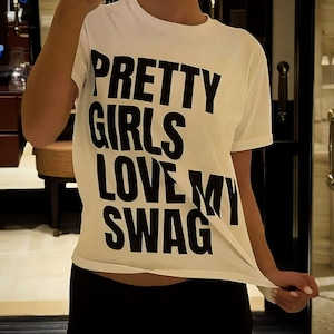 Puede incluir: Una camiseta blanca de manga corta con el texto negro PRETTY GIRLS LOVE MY SWAG impreso en una fuente negrita y sin remates en el pecho. La prenda tiene un cuello redondo y un corte estándar, combinada con unos pantalones cortos negros.