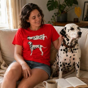 May include: A bright red short-sleeved t-shirt featuring two graphic illustrations of a Dalmatian dog. One dog is depicted in a standing profile view, while the other is shown in a horizontal position above it, both displaying black spots.
