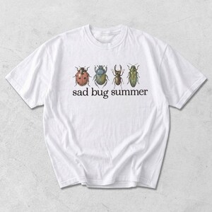 Pode incluir: Uma t-shirt branca com uma impressão gráfica de quatro insetos distintos dispostos numa linha horizontal acima do texto preto "sad bug summer". Os insetos incluem uma joaninha vermelha, um escaravelho azul, um escaravelho castanho e um besouro verde alongado.