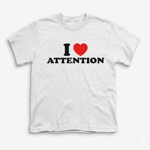 Op de afbeelding: Wit t-shirt met de tekst "I ❤️ ATTENTION" in zwart en rood. Het hartsymbool is rood en de tekst is in een vetgedrukte, schreefloze lettertype. Het shirt heeft korte mouwen en een ronde hals.