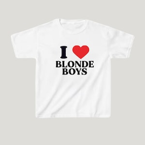 Op de afbeelding: Wit T-shirt met de tekst "I ❤️ BLONDE BOYS" in zwart en rood. Het ontwerp bevat een zwarte "I", een rood hartsymbool en de woorden "BLONDE BOYS" in een vet, schreefloos lettertype.