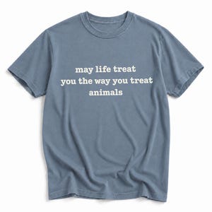 May include: A slate blue short-sleeve t-shirt featuring the white text "may life treat you the way you treat animals" printed across the center of the chest. The garment has a crew neckline and a standard fit with visible fabric texture.