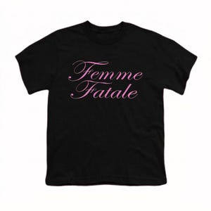 以下が含まれることがあります： 前面にピンク色の筆記体で「Femme Fatale」と書かれた黒いTシャツ。テキストは繊細な筆記体フォントです。シャツはクラシックなクルーネックです。