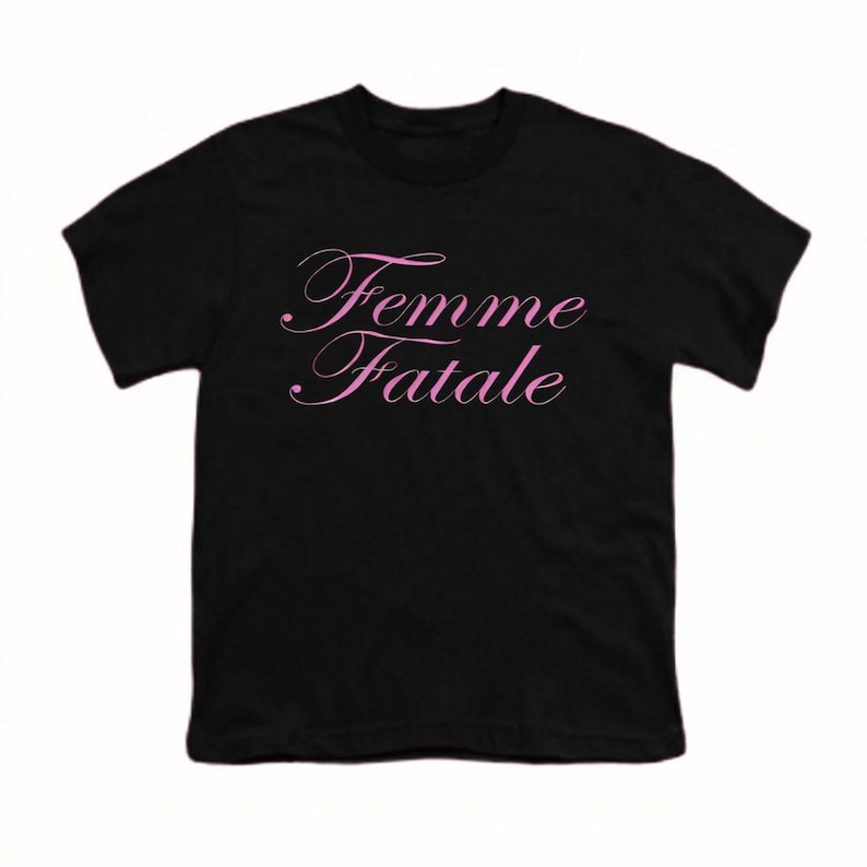 以下が含まれることがあります： 前面にピンク色の筆記体で「Femme Fatale」と書かれた黒いTシャツ。テキストは繊細な筆記体フォントです。シャツはクラシックなクルーネックです。
