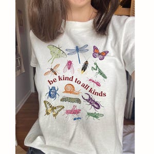Pode incluir: Uma t-shirt branca de manga curta com uma impressão gráfica colorida de vários insetos e um caracol. O texto "be kind to all kinds" está impresso a vermelho no centro. As ilustrações incluem borboletas, uma libélula, um escaravelho e uma formiga.