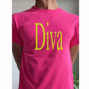 May include: A bright pink t-shirt with the word "Diva" printed in a contrasting yellow color. The text is centered on the front of the shirt. The shirt has short sleeves and a crew neck.