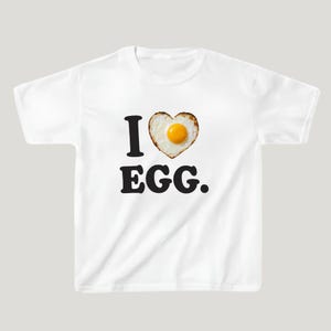 Könnte beinhalten: Ein weißes T-Shirt mit kurzen Ärmeln, das den schwarzen Text I, ein herzförmiges Spiegelei mit gelbem Eigelb und den schwarzen Text EGG zeigt. Die Grafik ist mittig auf der Brust platziert, das Shirt hat einen Rundhalsausschnitt.