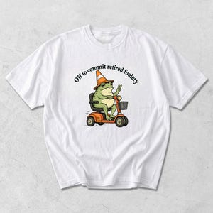 May include: A white t-shirt featuring a graphic of a green frog wearing an orange traffic cone hat, riding an orange mobility scooter, and making a peace sign. The text above the frog reads, Off to commit retired foolery in black font.