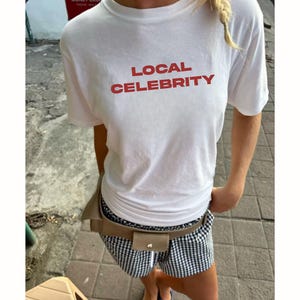 Może przedstawiać: Biały t-shirt z krótkim rękawem z czerwonym napisem LOCAL CELEBRITY na piersi. Koszulka jest zestawiona z czarno-białymi szortami w kratkę oraz beżową nerką noszoną w talii na wierzchu szortów.