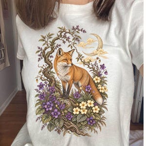 Puede incluir: Una camiseta blanca con un estampado gráfico detallado de un zorro rojo de pie entre un árbol nudoso, flores moradas y amarillas, y una luna creciente en el cielo. La ilustración está centrada en el pecho y utiliza tonos tierra.