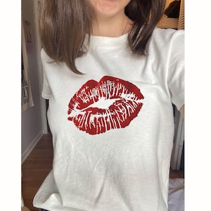 Könnte beinhalten: Weißes T-Shirt mit einem großen roten Lippenstift-Kuss-Aufdruck. Die Grafik befindet sich mittig auf der Vorderseite des Shirts. Das Shirt besteht aus einem weichen, hellen Stoff.