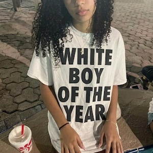 May include: A white short-sleeved t-shirt featuring the bold black text "WHITE BOY OF THE YEAR" printed in a stacked, sans-serif font across the chest. The garment has a classic crew neckline and a relaxed fit, made of a plain white cotton fabric.