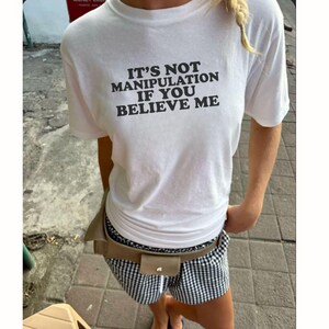 Może przedstawiać: Biały t-shirt z krótkim rękawem z czarnym napisem IT'S NOT MANIPULATION IF YOU BELIEVE ME na piersi, zestawiony z czarno-białymi szortami w kratkę vichy oraz beżową nerką noszoną w talii.