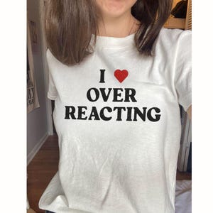 May include: A white short-sleeved t-shirt featuring the black text "I" followed by a red heart symbol and the words "OVER REACTING" printed in a bold, distressed font across the center of the chest area of the garment.