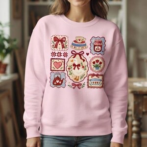 May include: A light pink crewneck sweatshirt featuring a central graphic print of a white goose surrounded by various small, framed illustrations including a red bow, a teapot, a heart, a jar of jam, tulips, and a baking dish with red accents.