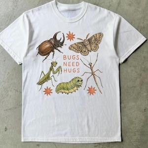 May include: A white short-sleeve t-shirt featuring a central graphic of a brown beetle, a moth, a green praying mantis, a green caterpillar, and a stick insect. The text "BUGS NEED HUGS" is printed in orange letters, surrounded by small orange star shapes.