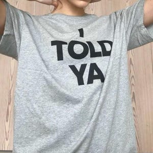 Könnte beinhalten: Ein hellgraues T-Shirt mit dem Text "I TOLD YA" in großen, schwarzen Buchstaben. Das T-Shirt hat kurze Ärmel und eine lockere Passform. Das Bild zeigt das T-Shirt an einer Person.