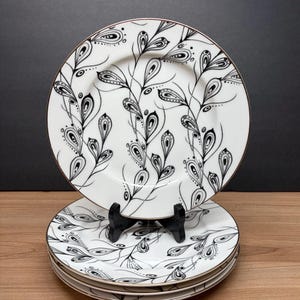 Peut inclure: Ensemble d'assiettes en c&eacute;ramique blanche avec un motif floral noir et blanc. Les assiettes ont un bord dor&eacute; et sont empil&eacute;es sur une surface en bois. Les assiettes sont pr&eacute;sent&eacute;es sur un support noir sur fond sombre.