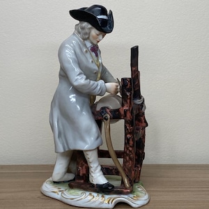 May include: A porcelain figurine depicting a man in a long gray coat and black hat, working at a printing press. The figurine has a detailed design with gold accents on the base and is set on a wooden surface.