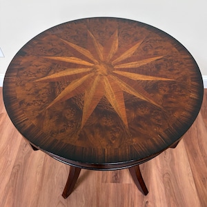 May include: A round wooden table featuring a dark brown burl wood veneer surface with a prominent light brown starburst inlay pattern at the center. The table has a smooth, polished finish and is supported by curved wooden legs extending downward.