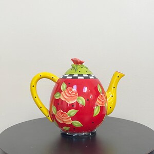 May include: A decorative teapot with a red body adorned with rose illustrations. The teapot features a yellow handle and spout with black polka dots, a green lid with a red rose, and a black and white checkered pattern on the rim.