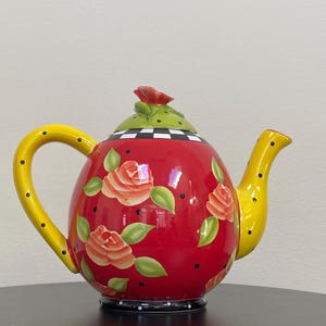 May include: A decorative red ceramic teapot with a yellow handle and spout. The teapot is adorned with orange roses, green leaves, and black polka dots. The lid is green with a red rose and a black and white checkered pattern.
