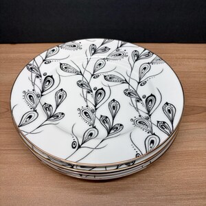 Peut inclure: Une pile de quatre assiettes en c&eacute;ramique blanche avec un motif floral noir et blanc. Les assiettes ont un bord dor&eacute;. Le motif floral pr&eacute;sente des feuilles et des branches stylis&eacute;es. Les assiettes sont empil&eacute;es sur une surface en bois.