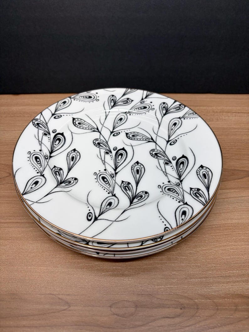 Peut inclure: Une pile de quatre assiettes en c&eacute;ramique blanche avec un motif floral noir et blanc. Les assiettes ont un bord dor&eacute;. Le motif floral pr&eacute;sente des feuilles et des branches stylis&eacute;es. Les assiettes sont empil&eacute;es sur une surface en bois.
